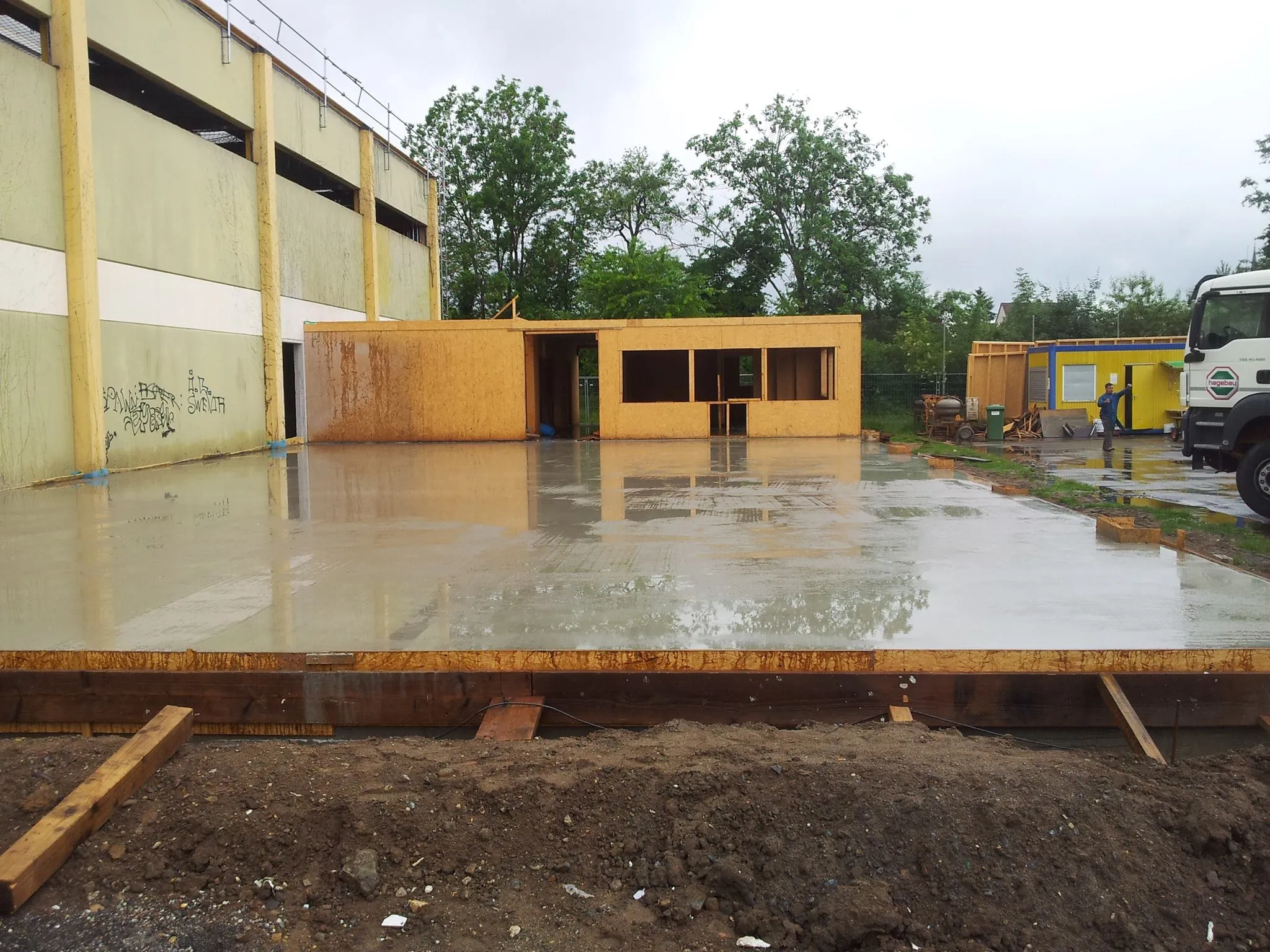 Frisch gegossene Betonbodenplatte im Regen, dahinter der begonnene Holzrohbau und ein Lastwagen