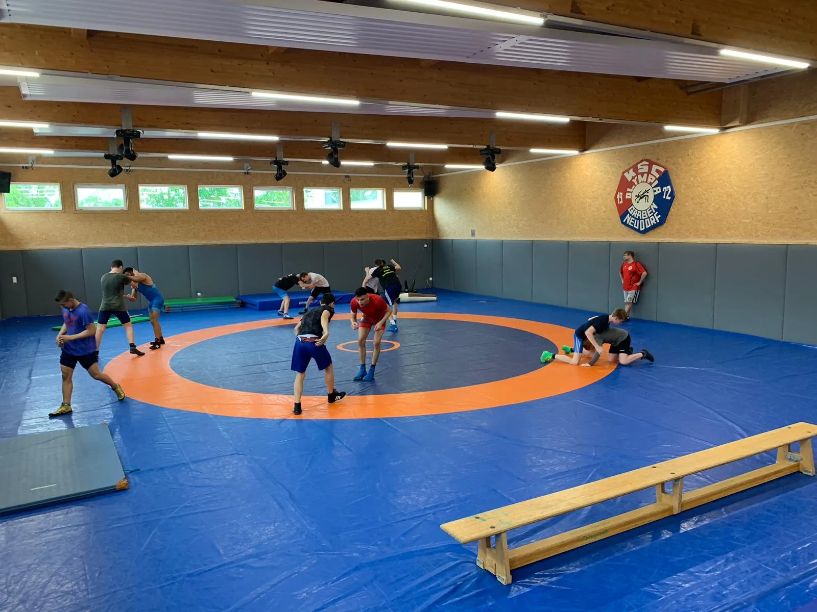 Blick in die Ringerhalle des Vereins: Wettkampfmatte mit orangefarbenem Kampfkreis, das Vereinswappen an der Wand, etwa ein Dutzend Ringer im Training