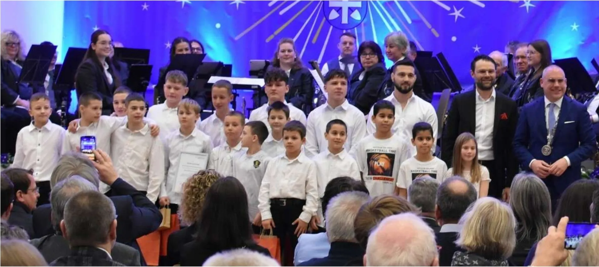Ehrung beim Neujahrsempfang der Gemeinde: die Mannschaft in weißen Hemden auf der Bühne, rechts der Bürgermeister mit Amtskette, im Hintergrund ein Orchester